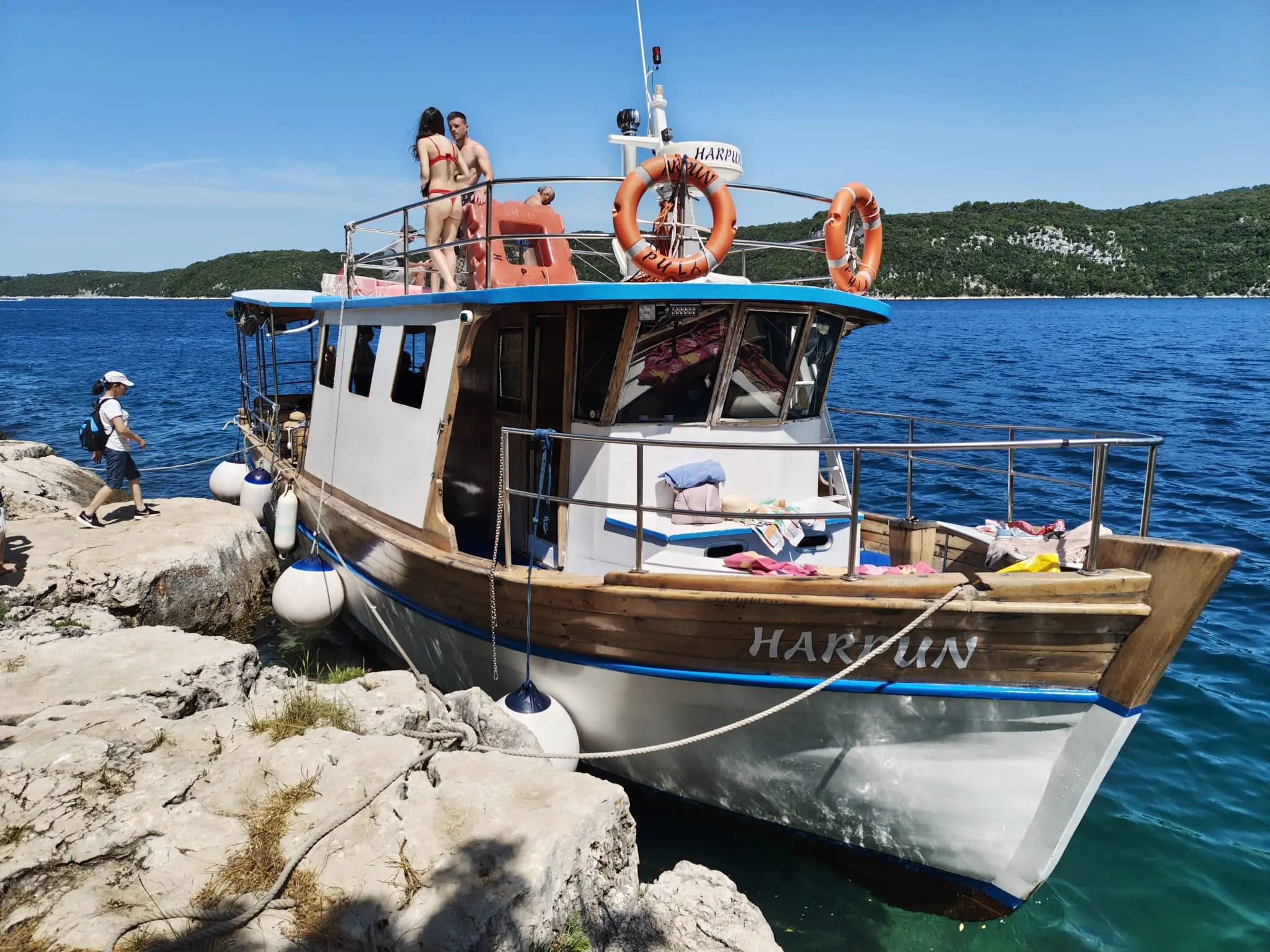 The Harpun boat moored in a rocky cove, guests on the sundeck
